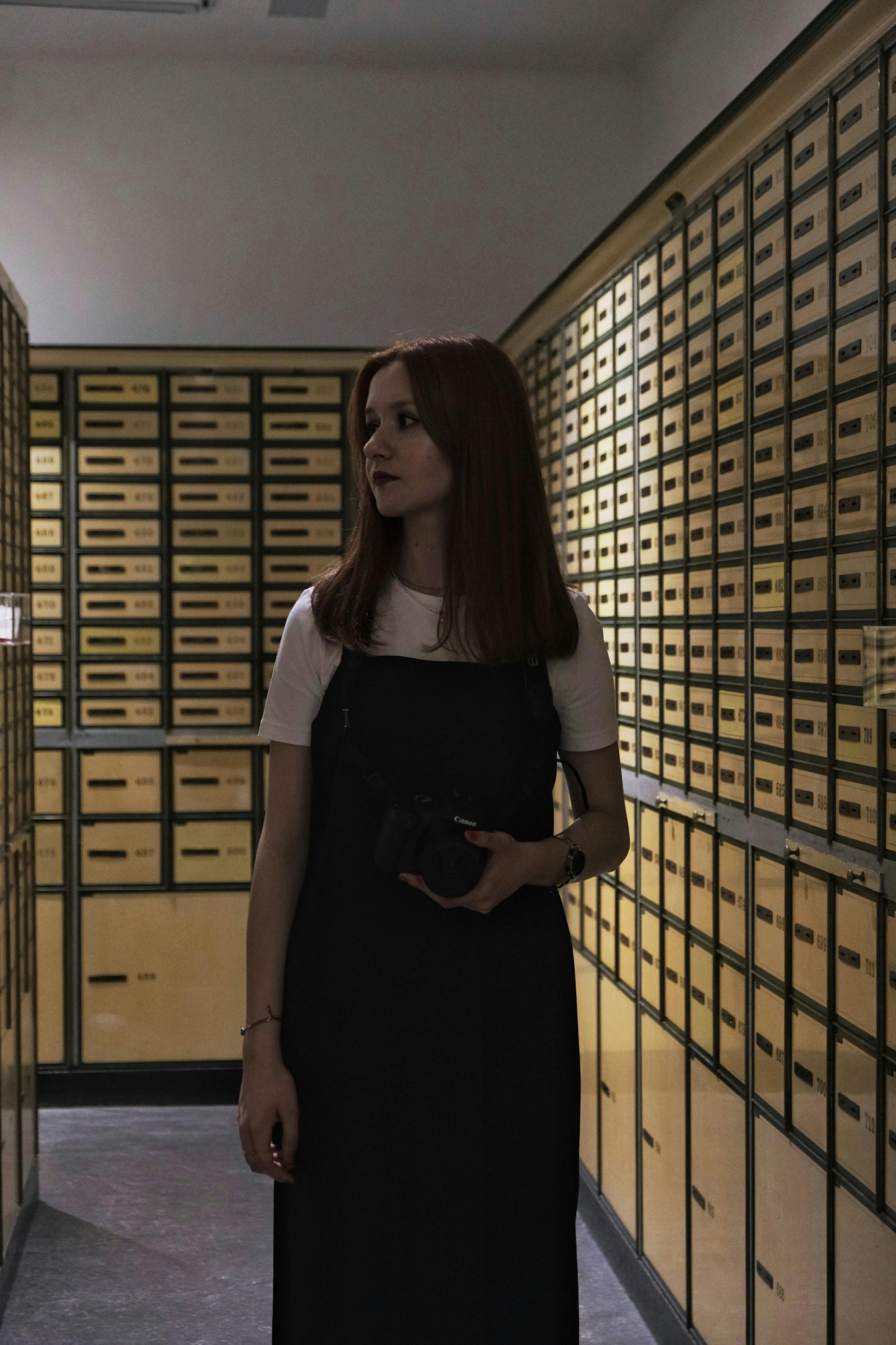 Young woman standing in a vault surrounded by safety deposit boxes, holding a camera.