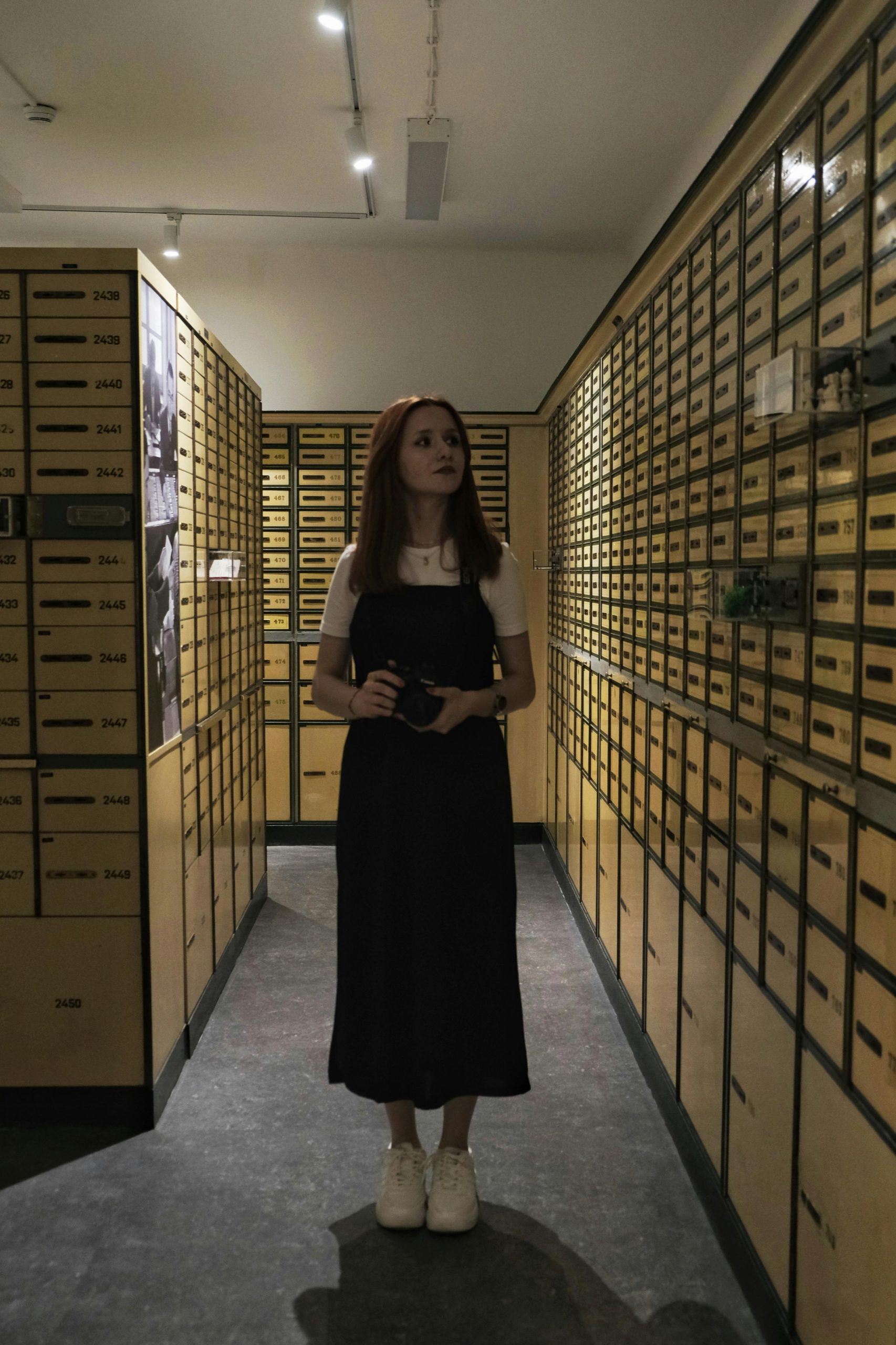 Young woman standing in a vault surrounded by safety deposit boxes, holding a camera.